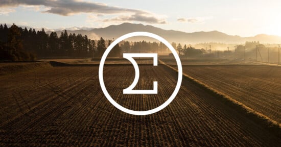 A plowed field at sunrise with mountains in the background, trees lining the horizon, and a large white Sigma symbol enclosed in a circle overlaying the center of the image.