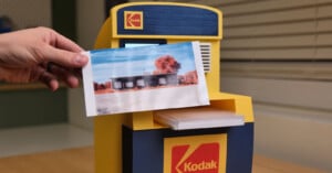 A hand holds a color photo in front of a small yellow and blue Kodak-branded photo printer on a wooden table, with window blinds in the background.