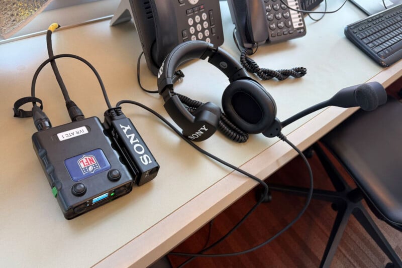 A Sony headset with a microphone is connected to an audio receiver labeled "NFL" and "WR ACT." The equipment is on an office desk with two office phones, a keyboard, and a chair nearby.