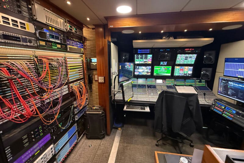 A broadcast control room filled with racks of cables and equipment on the left, and multiple monitors, audio mixers, and computers on desks to the right. The room is brightly lit and organized for live production.