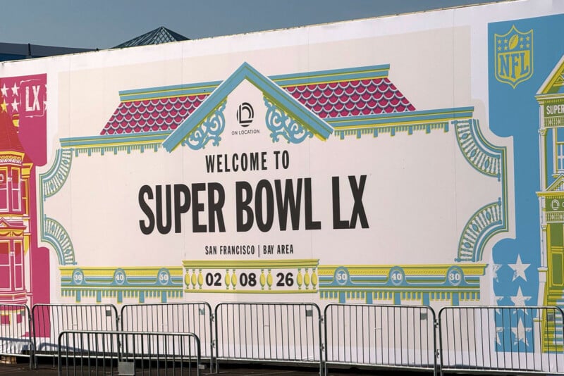 A large sign reads "Welcome to Super Bowl LX, San Francisco | Bay Area, 02.08.26" with colorful house designs and NFL logos, behind metal barricades.