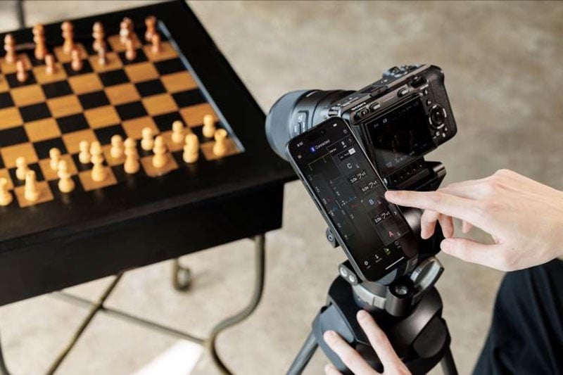 Una persona usa un teléfono inteligente montado en un trípode de cámara para grabar o transmitir un tablero configurado para un juego, con las piezas dispuestas y la cámara apuntando al tablero.