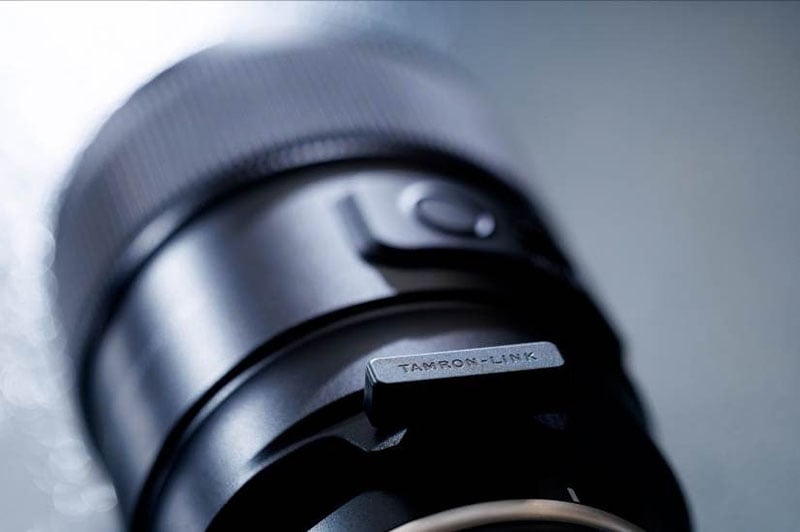 Primer plano de la lente de la cámara con foco en el interruptor de metal marcado "ENLACE Tamron," Muestra detalles finos y fondos borrosos.