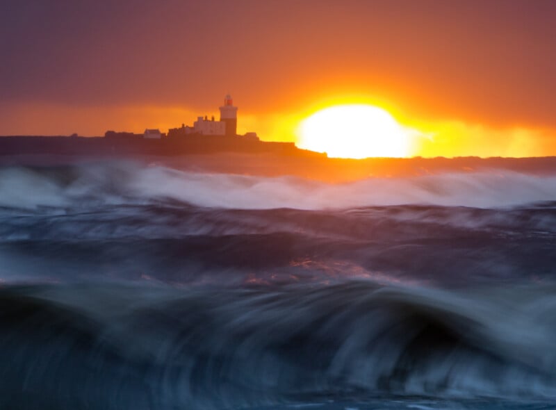 Waves crash in the foreground as the sun sets dramatically behind a distant lighthouse on the coastline, casting a warm orange and yellow glow across the sky and water.