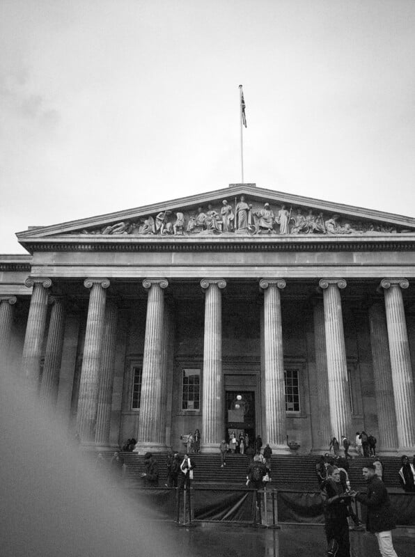 Black and white photo of the British Museum’s neoclassical facade, featuring tall columns, a pediment with sculptures, a flag on top, and people gathered at the entrance on a cloudy day.