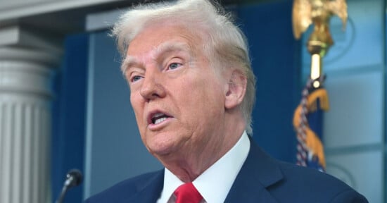 A man with light skin and blond hair, wearing a dark suit, white shirt, and red tie, speaks in front of a podium. An American flag and a gold eagle emblem are visible in the background.