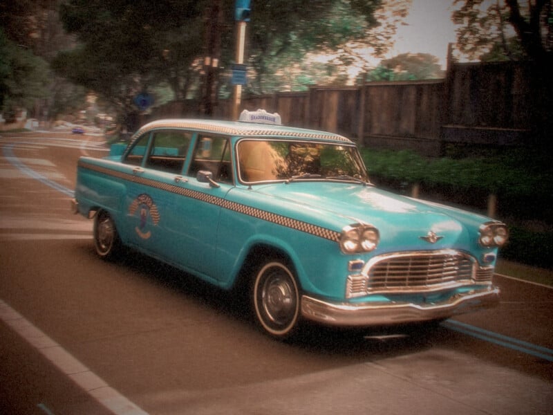 Un antiguo taxi azul con decoración a cuadros conduce por una calle arbolada de la ciudad en un día nublado. Esta foto tiene un filtro suave y nostálgico.
