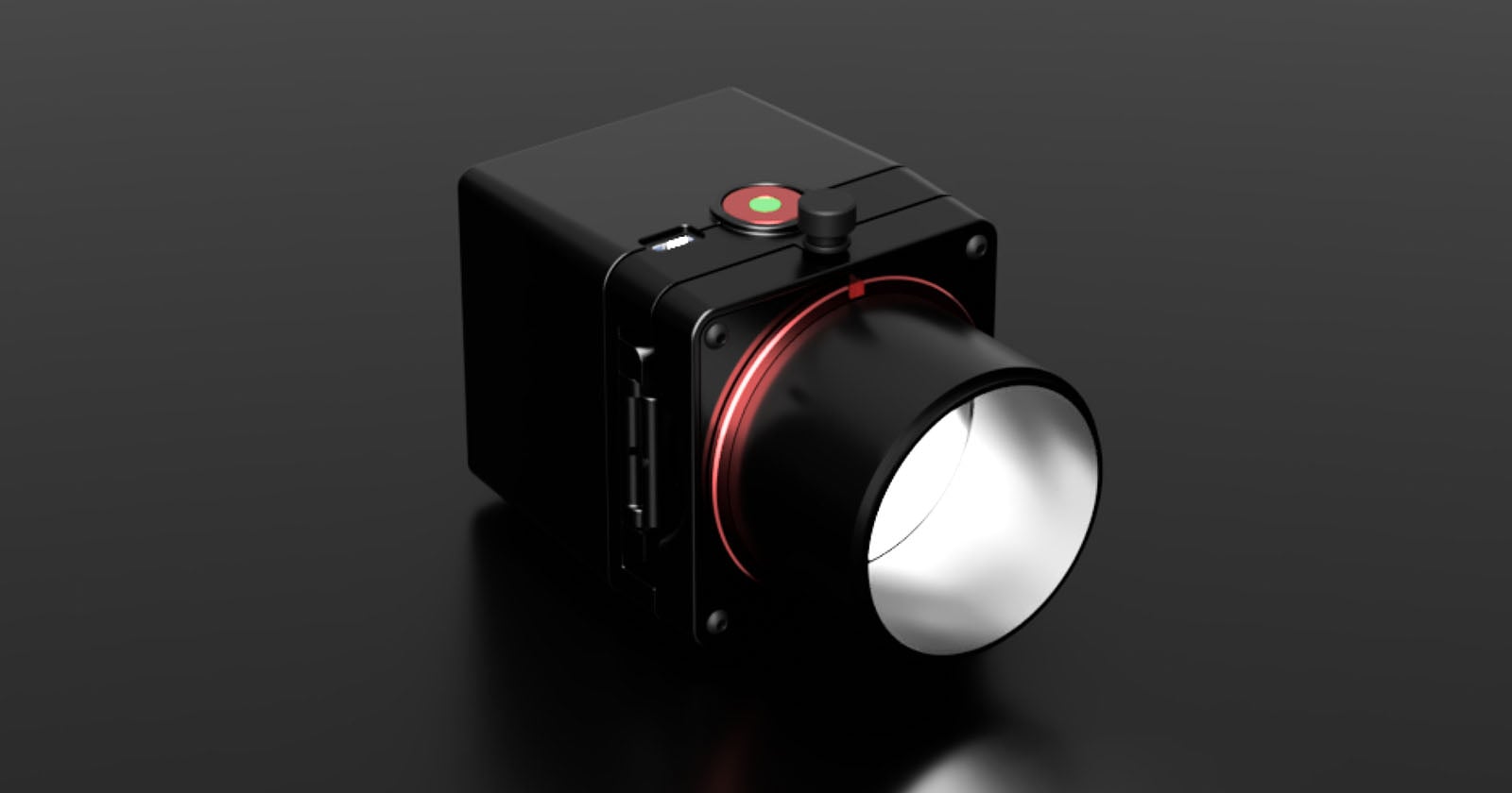 A black, box-shaped camera with a large lens and a red ring around its base sits on a reflective dark surface. The camera has a round green and red button on top and several ports on the side.