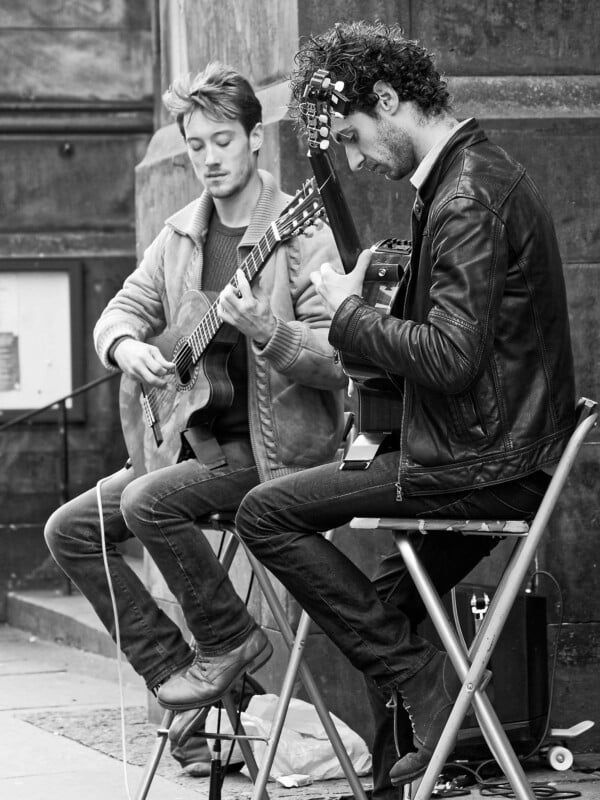 Dos hombres tocando la guitarra acústica mientras estaban sentados en taburetes en una calle de la ciudad. Uno lleva una chaqueta de cuero y el otro un jersey. Ambos se concentraron en sus instrumentos, tocando música juntos al aire libre. La imagen está en blanco y negro.