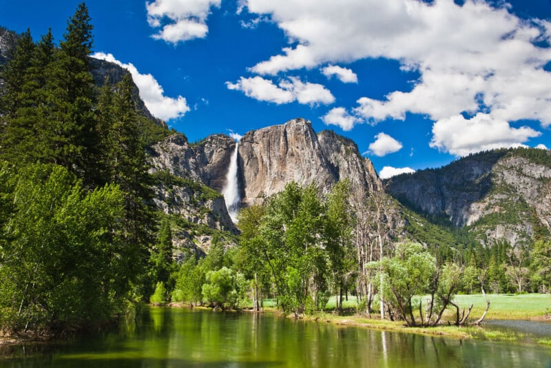 A tall waterfall cascades down a rocky cliff surrounded by green trees and a calm river under a bright blue sky with scattered white clouds in a mountain landscape.