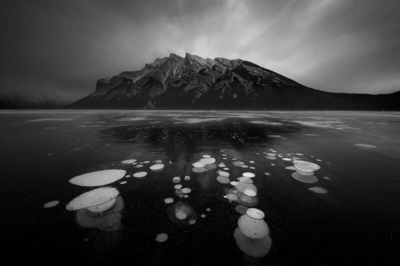 A dramatic black-and-white photo of a frozen lake with circular ice bubbles beneath the surface, set against rugged mountains under a moody, cloudy sky.