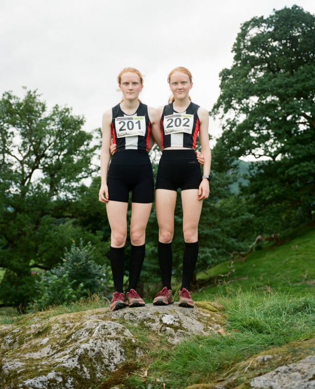 Two young women with similar appearance, wearing matching athletic outfits and numbered bibs 201 and 202, stand side by side on a rock outdoors, surrounded by green trees and grass.