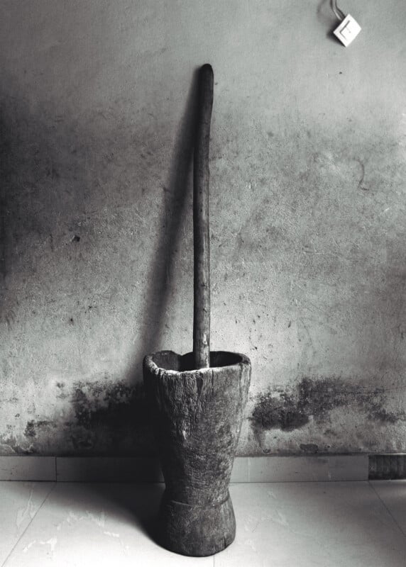 A traditional wooden mortar and pestle stands against a weathered, textured wall; an electrical socket is visible in the top right corner. The floor is tiled and the image is in black and white.