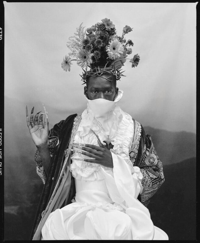 A person in elaborate, ornate clothing and a flowing robe sits against a neutral backdrop. They wear a floral crown, long false nails, and a face covering, with an expressive, regal pose and intense gaze. The photo is in black and white.