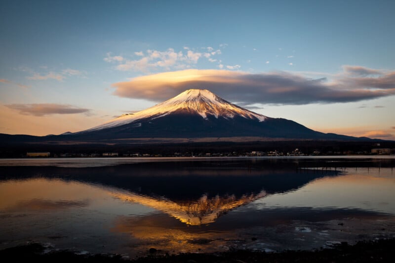Snow-capped Mount Fuji at sunrise, with clouds above and its reflection visible in a calm lake below. The sky is mostly clear with soft, warm lighting.