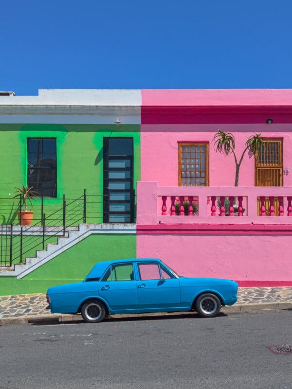 A bright blue vintage car parked in front of a building painted green on one side and pink on the other, with clear blue sky overhead and two potted plants near the entrance.