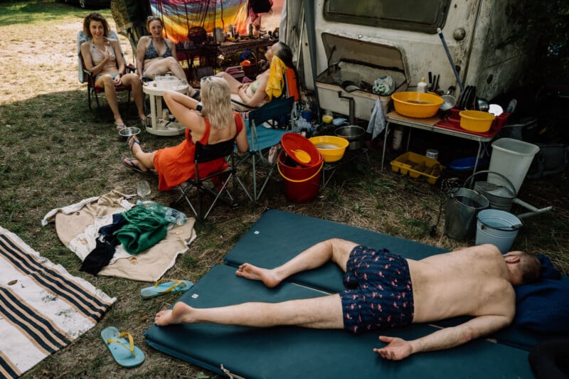 A man lies face up on a mat in swim trunks, while four people sit and talk near a camper with colorful bowls, towels, and buckets scattered around outdoors on a sunny day.