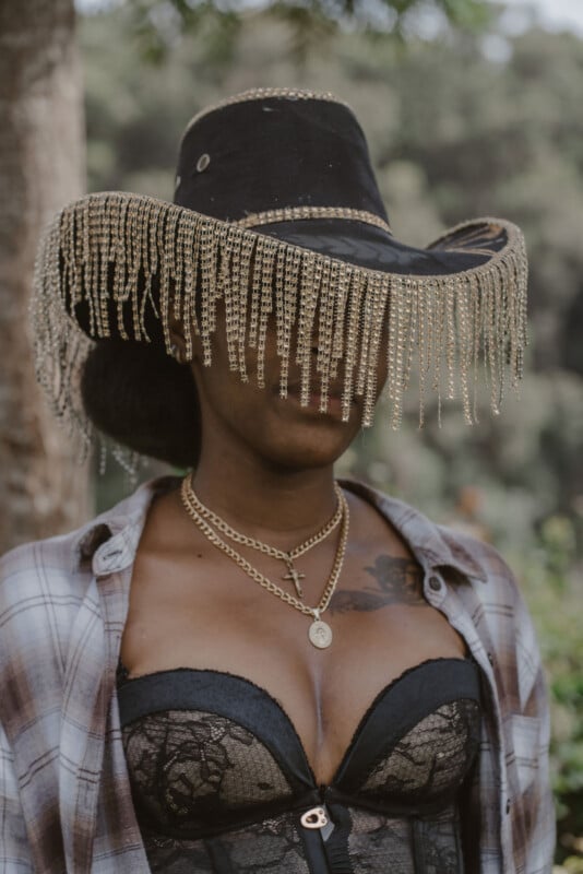 A woman wearing a black cowboy hat with gold fringe covering her eyes, a plaid shirt, layered gold necklaces, and a black lace bustier stands outdoors with trees in the background.