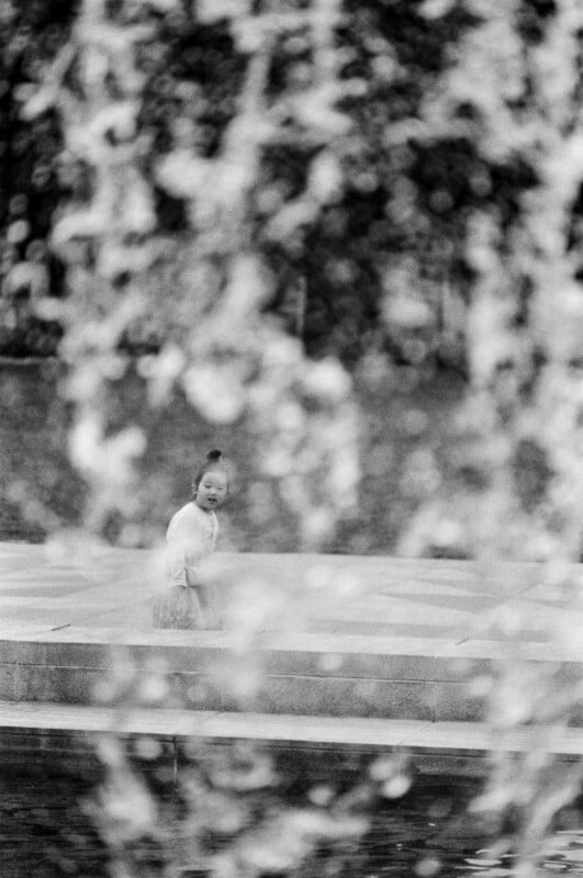 A black and white photo of a young child standing by a fountain, partially obscured by streams of water in the foreground, with a blurred background of trees and pavement.