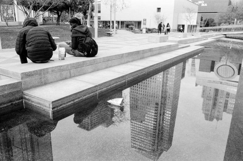 Two people sit on a concrete ledge near a shallow reflecting pool in an urban plaza, with city buildings and trees visible and reflected in the water; a paper boat floats in the pool.