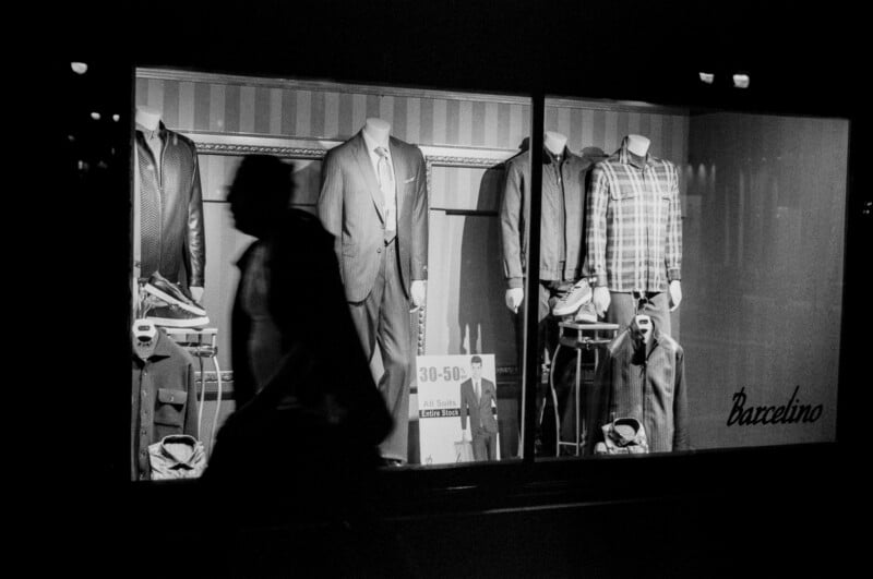 A silhouette of a person walks past a brightly lit store window displaying mannequins dressed in men's suits, shirts, and jackets. A sign in the window advertises discounts of 30–50%.