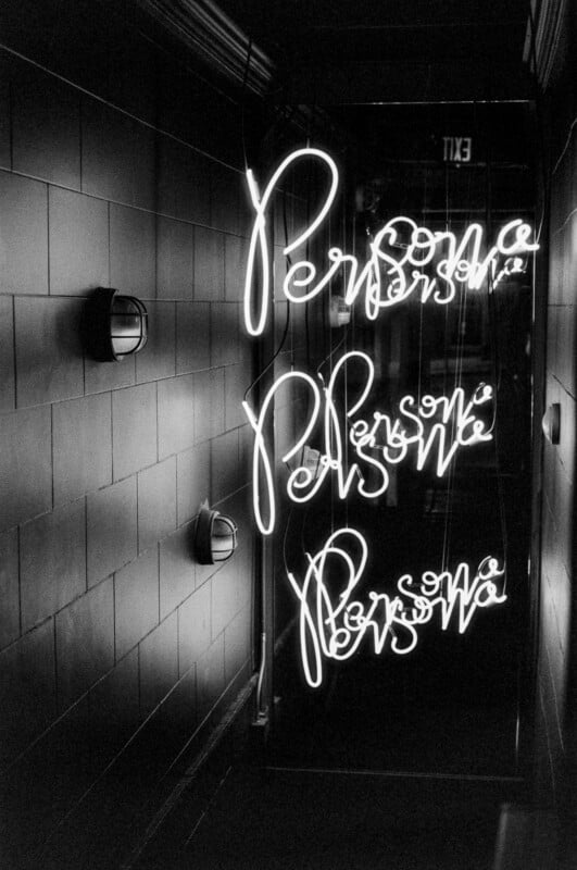 A black-and-white photo showing the word "Persona" written in neon lights, reflected multiple times in a mirror along a tiled wall in a dimly lit hallway.