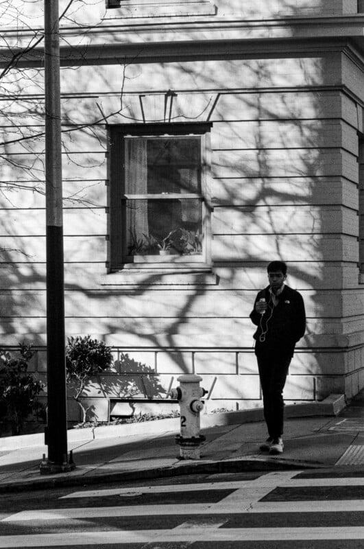 A man in dark clothing stands on a city sidewalk near a fire hydrant, looking at his phone with earphones in. Shadows of tree branches are cast on the building wall behind him.