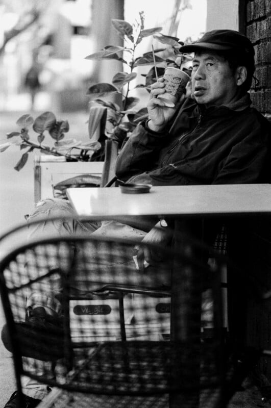 A man in a cap sits at an outdoor café table, holding a paper cup and looking to the side. Plants and another empty chair are visible, with the scene in black and white.