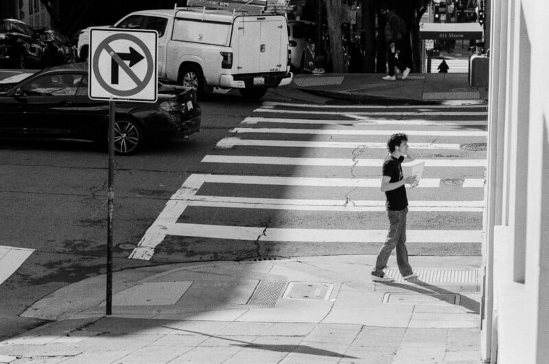 A person walks across a city crosswalk holding papers, while a "No Left Turn" sign stands nearby. Cars are parked along the street, and sunlight casts a shadow over part of the scene.