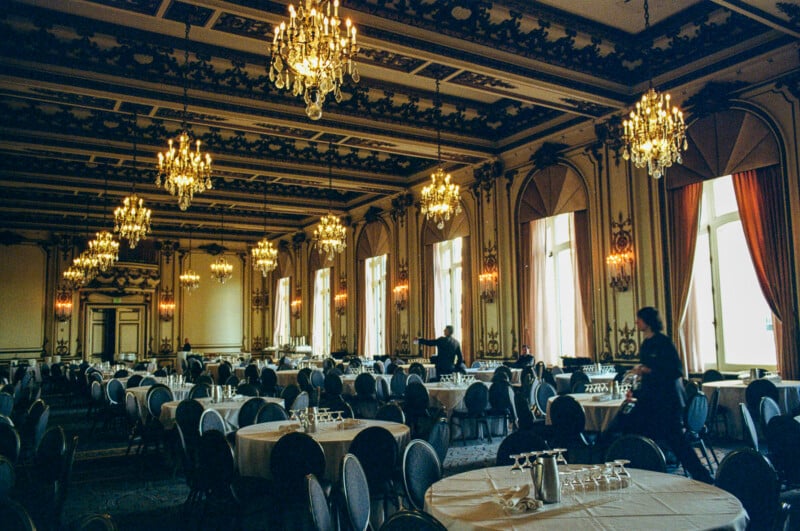 A grand banquet hall with chandeliers, high ornate ceilings, large windows with drapes, and round tables set with glasses. Several people are preparing or setting up the room.