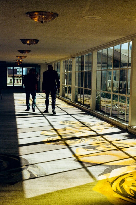 Two people walk down a sunlit hallway with large windows on the right. Shadows and patterns from the window frames stretch across the colorful carpet, creating an intricate design on the floor.