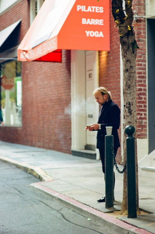 A man in a dark suit stands on a city sidewalk next to a tree, smoking a cigarette. Behind him is a brick building with an orange awning that reads “PILATES BARRE YOGA.” A clear plastic cup sits on a post nearby.