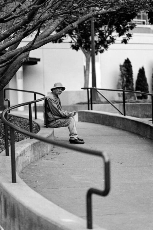 A man wearing a hat and jacket sits alone on a curved outdoor bench, looking towards the camera. Trees extend overhead, and a building with bushes is visible in the background. The photo is in black and white.