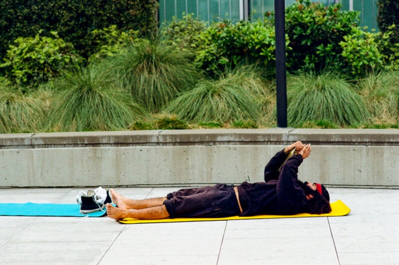 A person lies on their back on a yellow mat outdoors, holding their hands together above their chest. Their shoes and a bag are on the ground nearby, and green plants line the background.