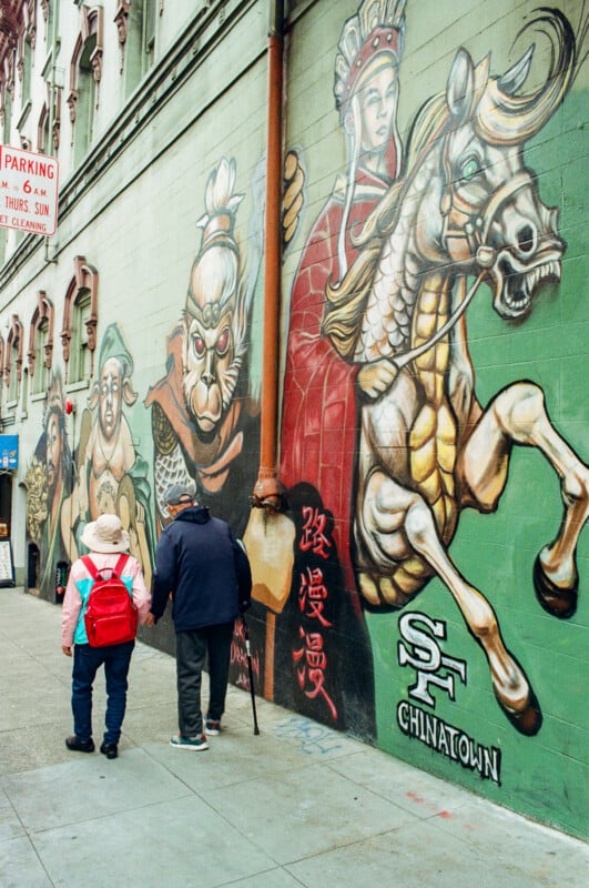 Two people walk past a large colorful mural in Chinatown, San Francisco, featuring warriors, a horse, and Chinese characters painted on a green wall.