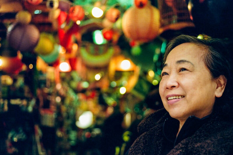 An older woman smiles while standing indoors, surrounded by colorful hanging lanterns and a softly-lit, festive background.