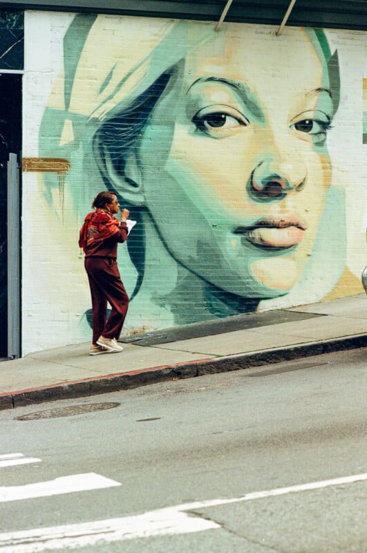 A woman wearing a red scarf and brown outfit walks uphill on a city sidewalk, sipping from a cup, in front of a large mural of a serene face painted on a wall.