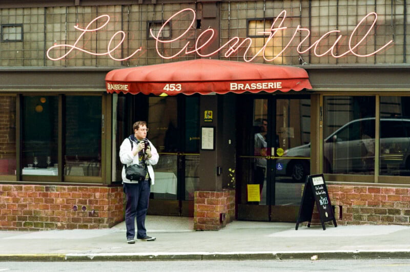 A person with a camera stands outside Le Central Brasserie under a red awning, with large cursive neon signage above and a sidewalk menu board in front. A reflection of a van is visible in the restaurant window.
