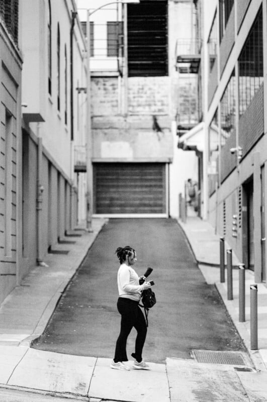 A woman stands on a sloped alleyway between tall buildings, holding a camera and looking to her left. The scene is in black and white, with strong urban and architectural elements.