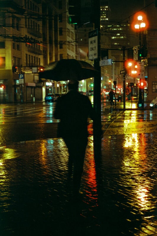 A person holding an umbrella walks alone on a wet city street at night, illuminated by colorful reflections from streetlights and traffic signals on the glistening pavement.