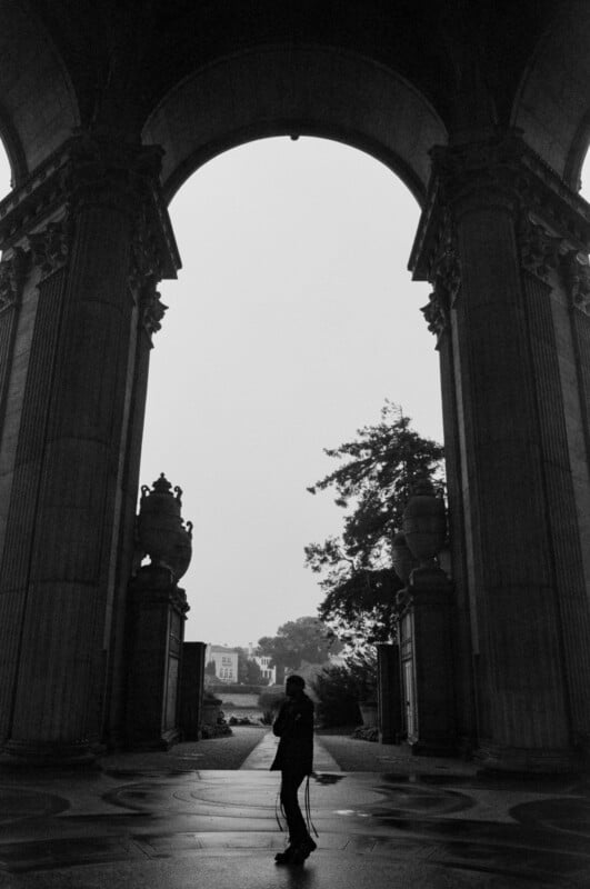 A silhouette of a person with a cane stands beneath a large archway with ornate columns, framed by trees and distant buildings under a cloudy sky.