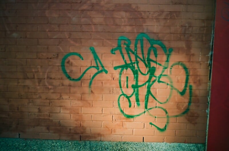 Green graffiti tag painted on a red brick wall, featuring overlapping and stylized letters that are difficult to read. Sunlight casts shadows on the wall and gravel is visible at the base.