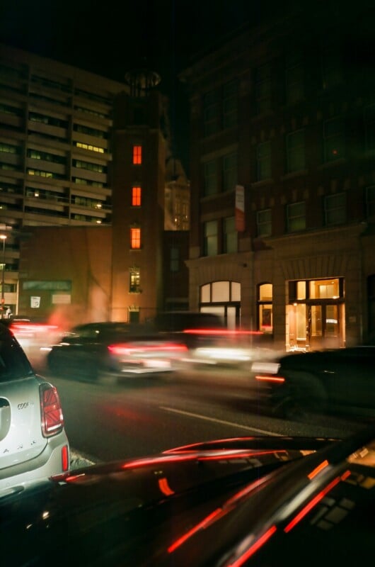 Cars with blurred headlights drive down a city street at night. Lights glow from windows of surrounding buildings, and a parked car is visible in the foreground. The scene is urban and dimly lit.