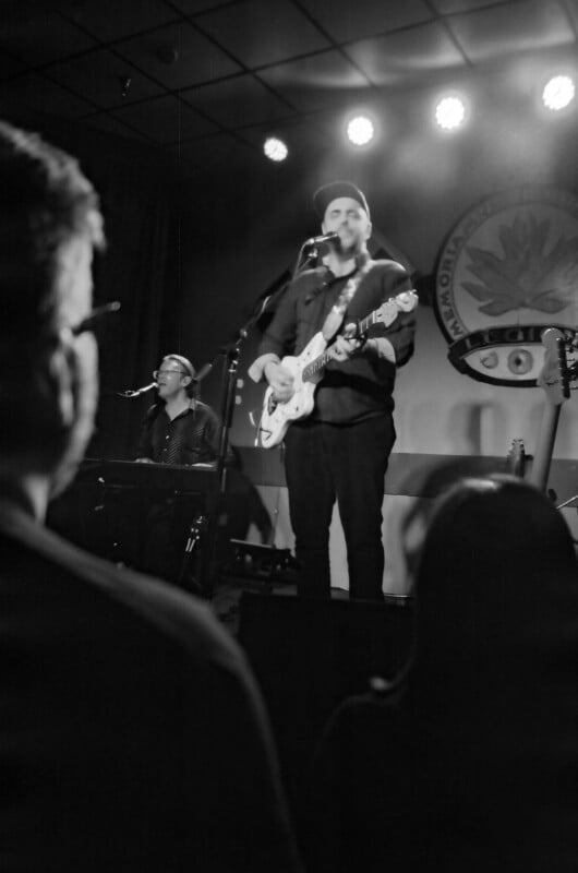 Black and white photo of a band performing on stage. A man in a cap plays electric guitar and sings into a microphone, while another musician plays keyboard. Two audience members are visible in the foreground.