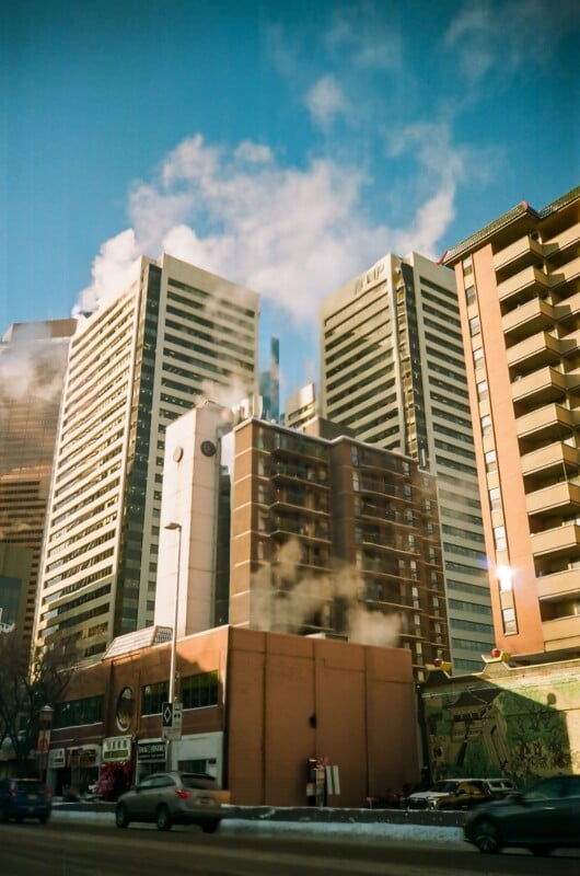 Tall office buildings with steam rising from rooftops loom over a busy city street with cars driving by; the sky is clear and blue, and some snow is visible on the ground and building ledges.