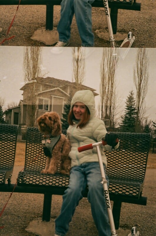 A young girl in a light jacket sits on a park bench holding a scooter, smiling next to a small brown dog. Leafless trees and a house are visible in the background on a cloudy day.