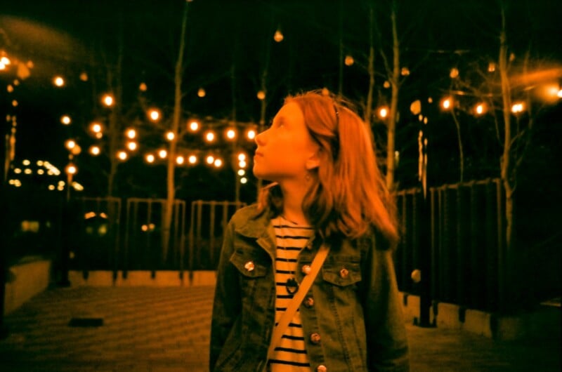 A young girl with shoulder-length hair and a striped shirt stands outside at night, looking up at string lights glowing warmly above her, creating a cozy and dreamy atmosphere.
