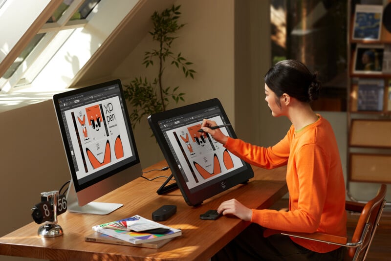A person in an orange sweater uses a stylus on a drawing tablet, editing a graphic design displayed on two monitors at a wooden desk in a bright, modern workspace.