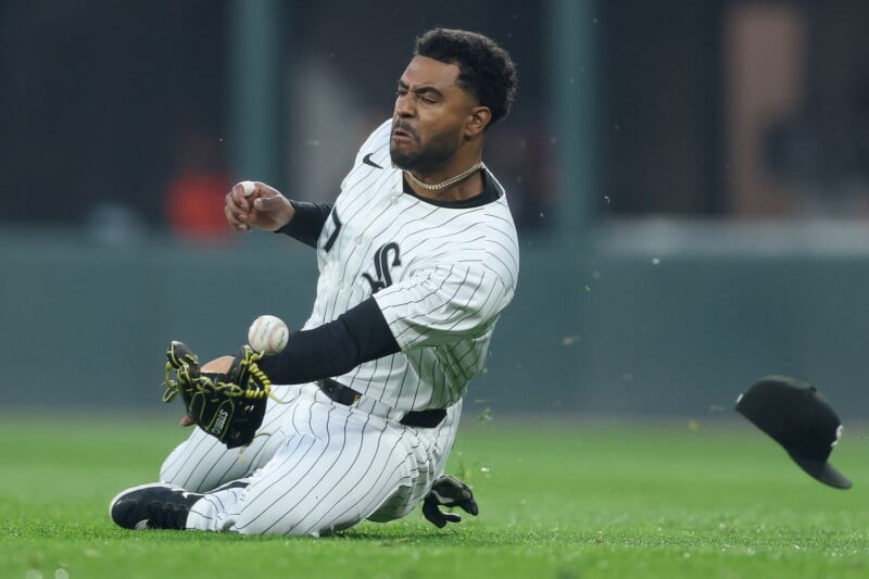 A baseball player in a white pinstripe uniform kneels on the grass, focused as he reaches to catch a baseball. His glove is extended, his hat has fallen off, and his expression shows determination.