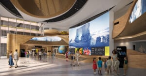 A spacious, modern museum lobby with large digital screens displaying nature scenes, wooden architectural features, a globe, informational exhibits, and visitors including adults and children walking and interacting.